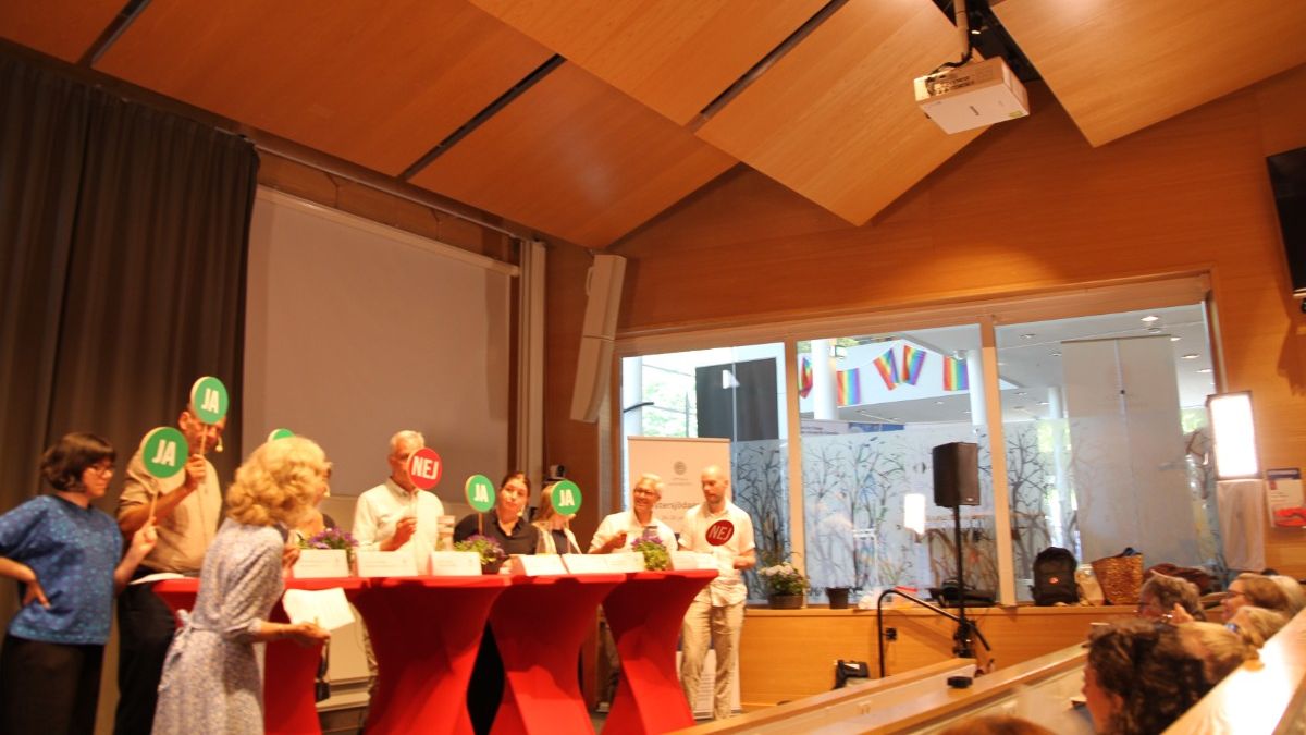 Politiker på scen i en lärosal. De lyfter gröna och röda ja/nej-skyltar framför publik.